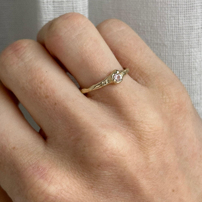 Hand wearing a gold ring with a pink gemstone against a neutral background