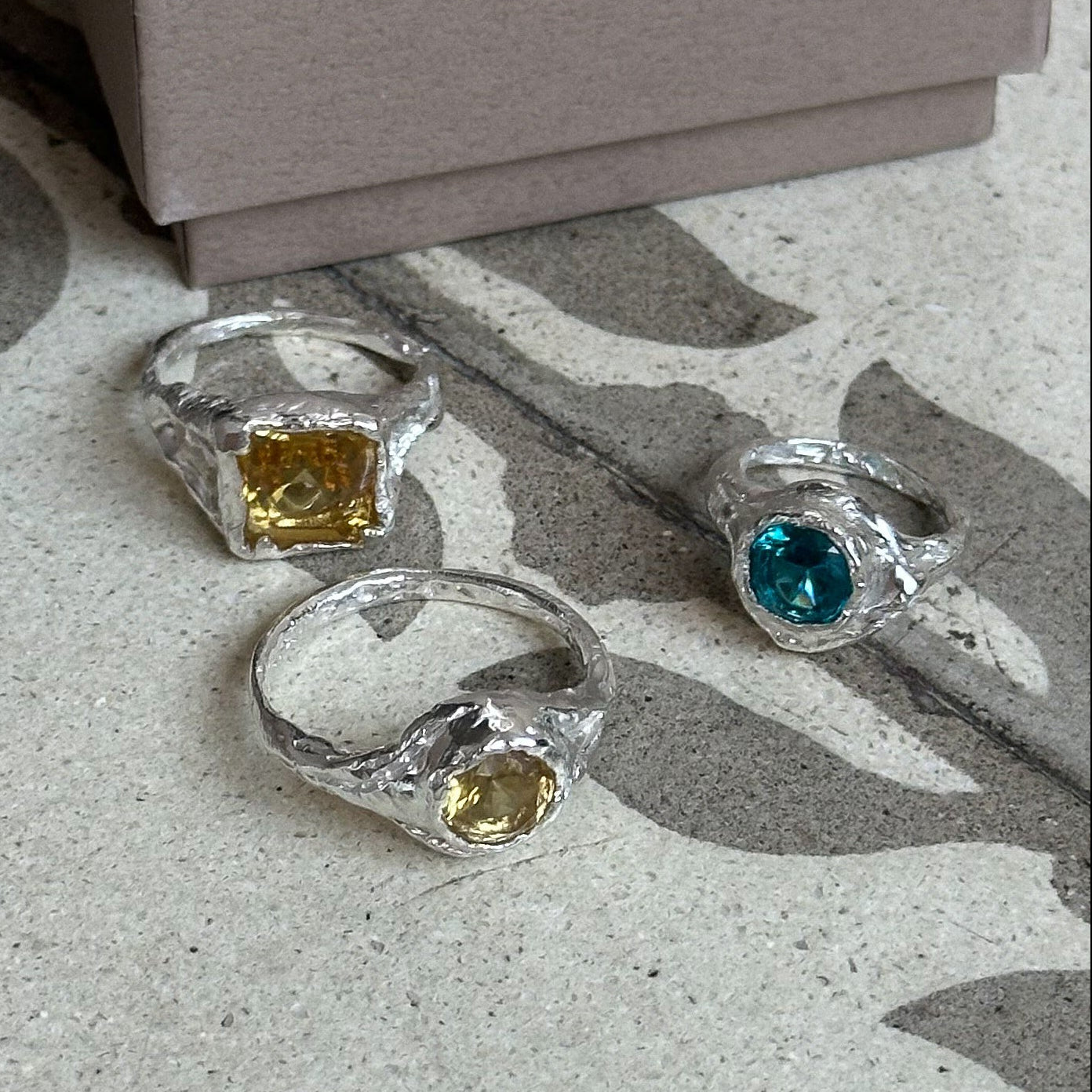 Silver rings with gemstones on a concrete surface next to branded boxes.