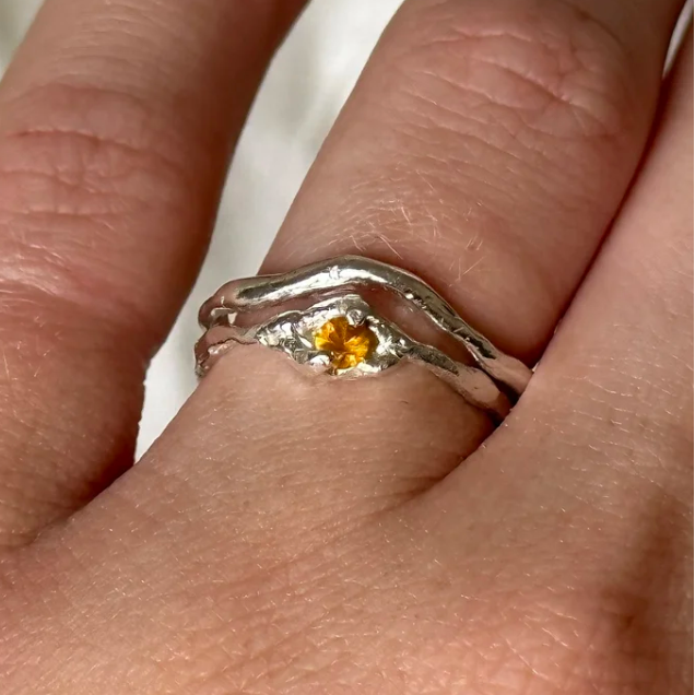 Silver ring with a yellow gemstone on a finger against a neutral background