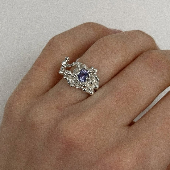 Silver ring with a purple gemstone on a hand against a plain background